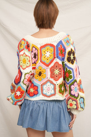 A person shown from the back wearing a colorful crocheted granny-square sweater and a light blue denim skirt against a light fabric backdrop.