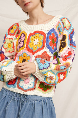 A close-up of a person wearing a colorful crocheted granny-square sweater with long puff sleeves and a cream base, paired with a light blue skirt against a soft off-white backdrop.