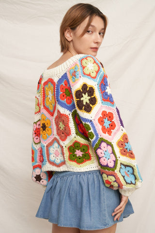 A woman wearing a colorful crocheted granny-square cardigan sweater and a light blue denim skirt, photographed against a neutral draped fabric backdrop.