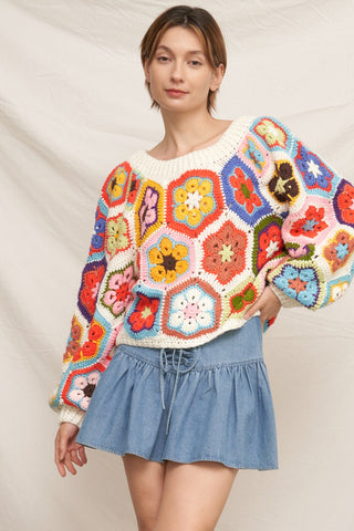 A woman wearing a colorful crocheted granny-square sweater and a light blue denim skirt, standing against a draped beige fabric backdrop.