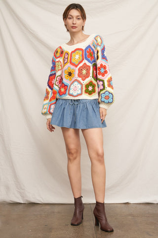 A woman modeling a cream-colored long-sleeve crochet sweater with colorful geometric hexagon patterns, paired with a blue denim mini skirt and brown ankle boots, standing against a draped off-white fabric backdrop.
