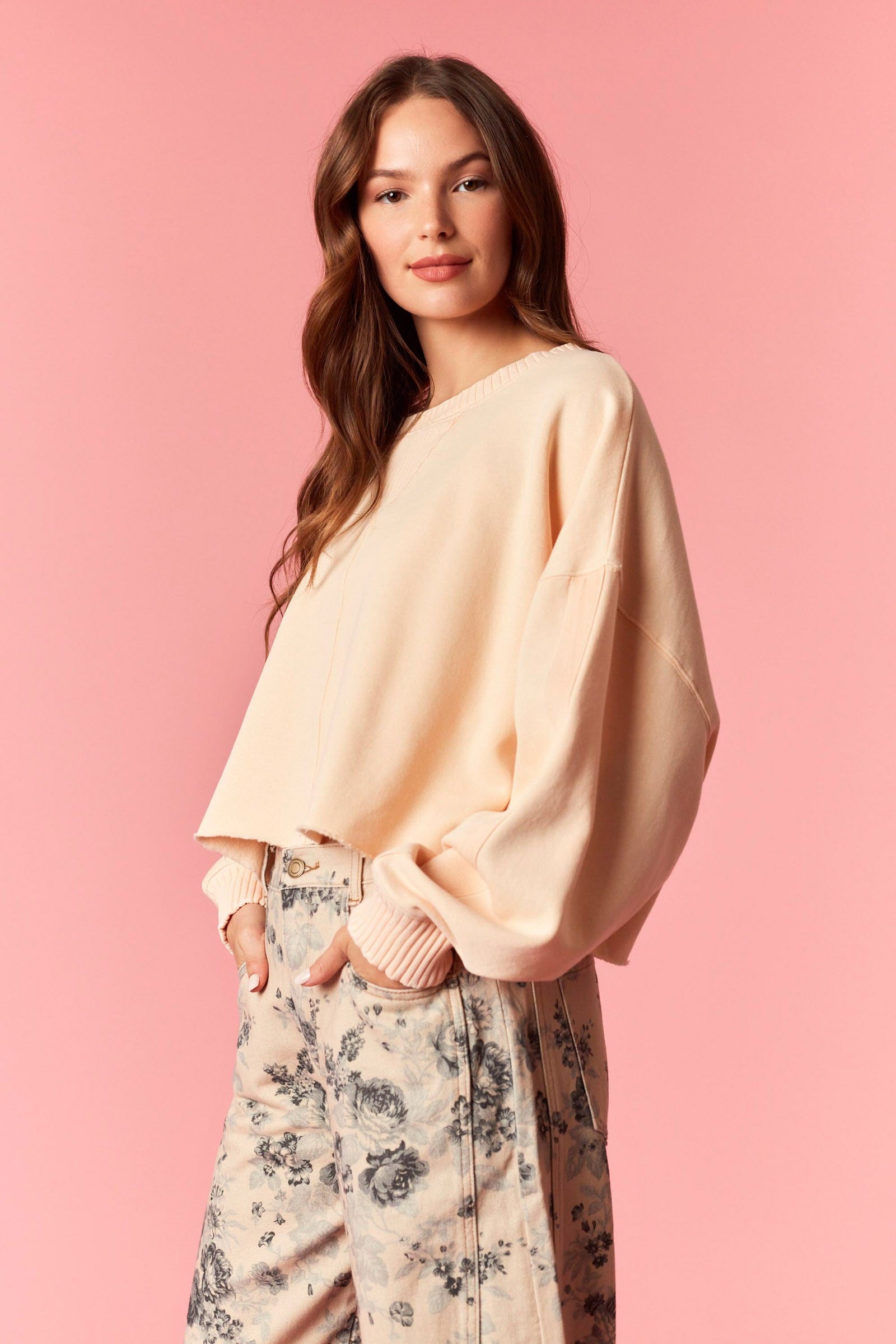 Woman wearing a light orange sweater and floral pants against a pink background
