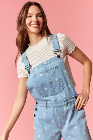 Woman wearing a white knit top and blue polka dot overalls on a pink background