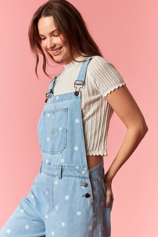 Woman wearing light blue denim overalls with white polka dots on a pink background