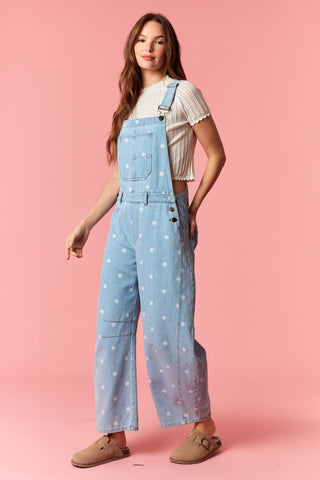 Woman wearing light blue denim overalls with white polka dots on a pink background