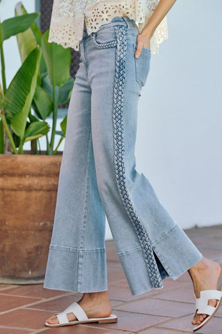 Person wearing light blue jeans with braided details, standing on a tiled floor with plants in the background.