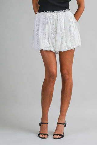 White lace ruffled shorts with a tie waist, worn by a model with black strappy high-heeled sandals against a light gray studio background.