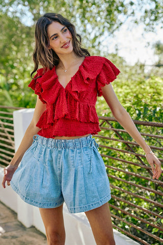 Woman wearing a red ruffled top and denim shorts standing outdoors with greenery in the background
