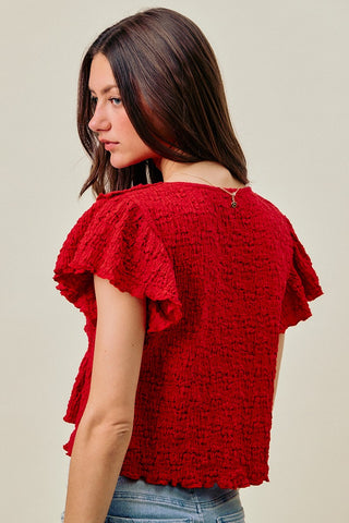 Woman wearing a red textured top with ruffled sleeves against a beige background