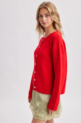 A woman modeling a bright red knit cardigan with white buttons, paired with a light beige skirt, against a plain light gray studio background.