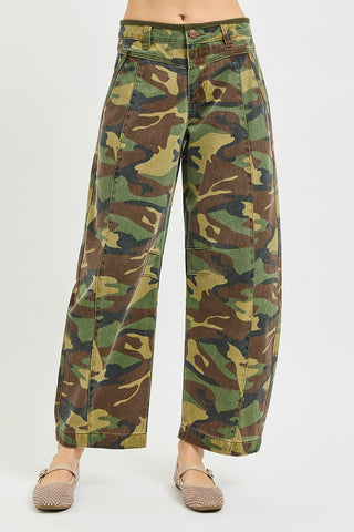 A person wearing high-waisted, wide-leg camouflage pants with a green, brown, and tan camo pattern, paired with brown flat shoes, shown from the waist down on a light gray background.