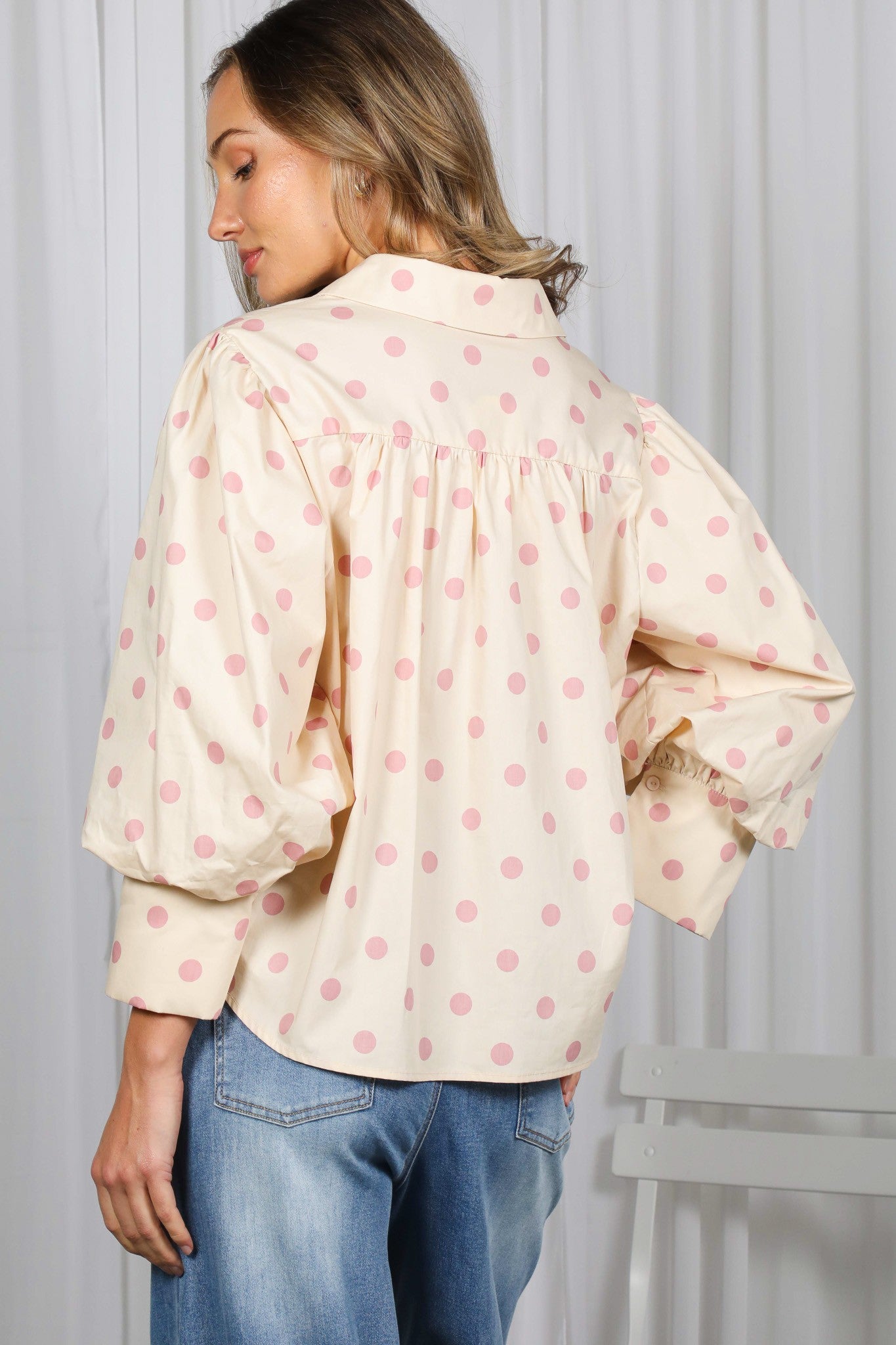 Woman wearing a cream blouse with pink polka dots and blue jeans against a white background