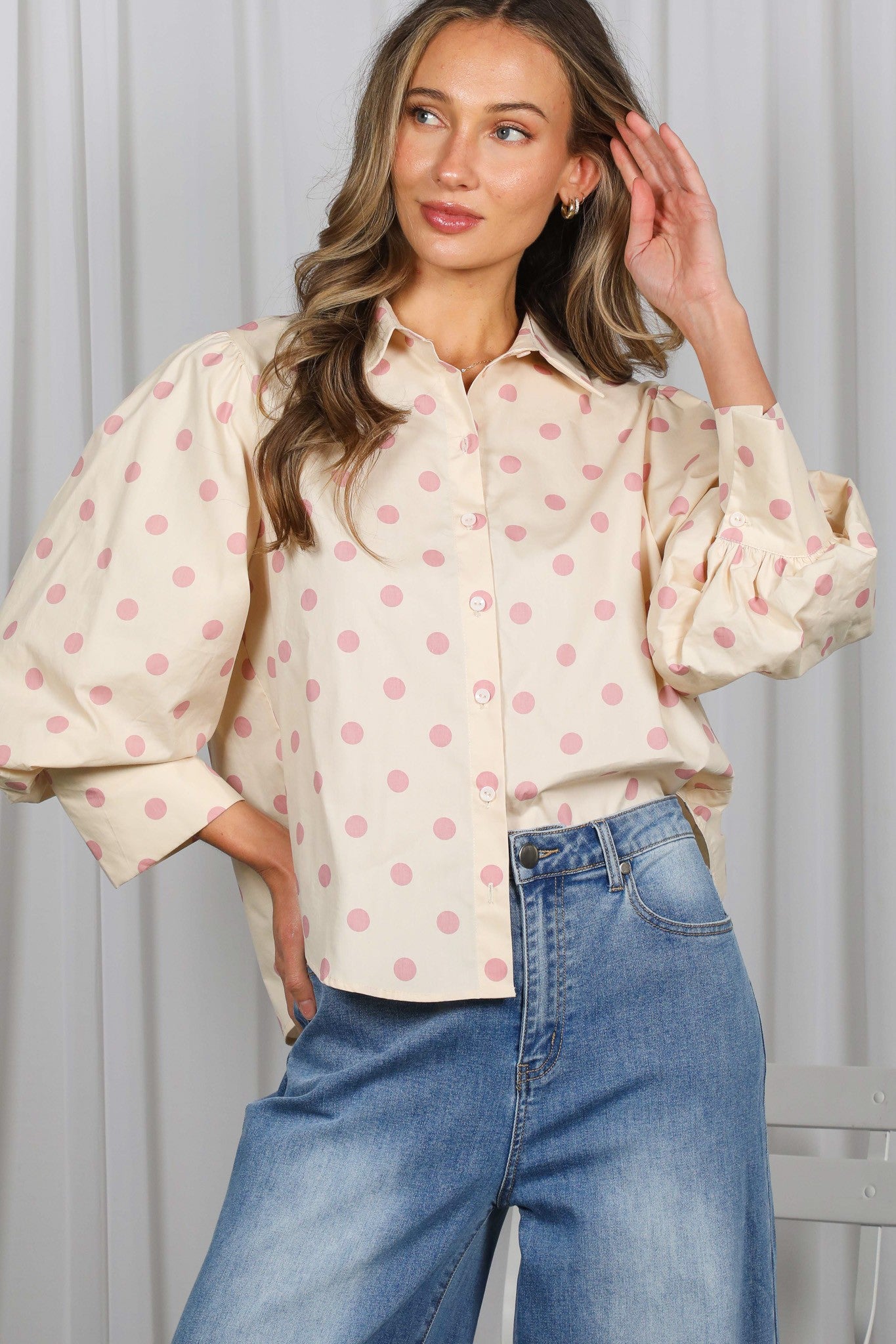 Woman wearing a cream blouse with pink polka dots and blue jeans against a white background