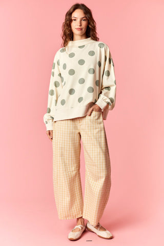 A woman stands against a solid pink studio background wearing a cream sweatshirt with sage green polka dots and beige-and-white gingham wide-leg pants.