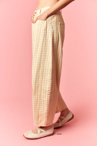 A person wearing wide-leg cropped pants in a beige and white gingham pattern, paired with white flat shoes, standing against a solid pink background.