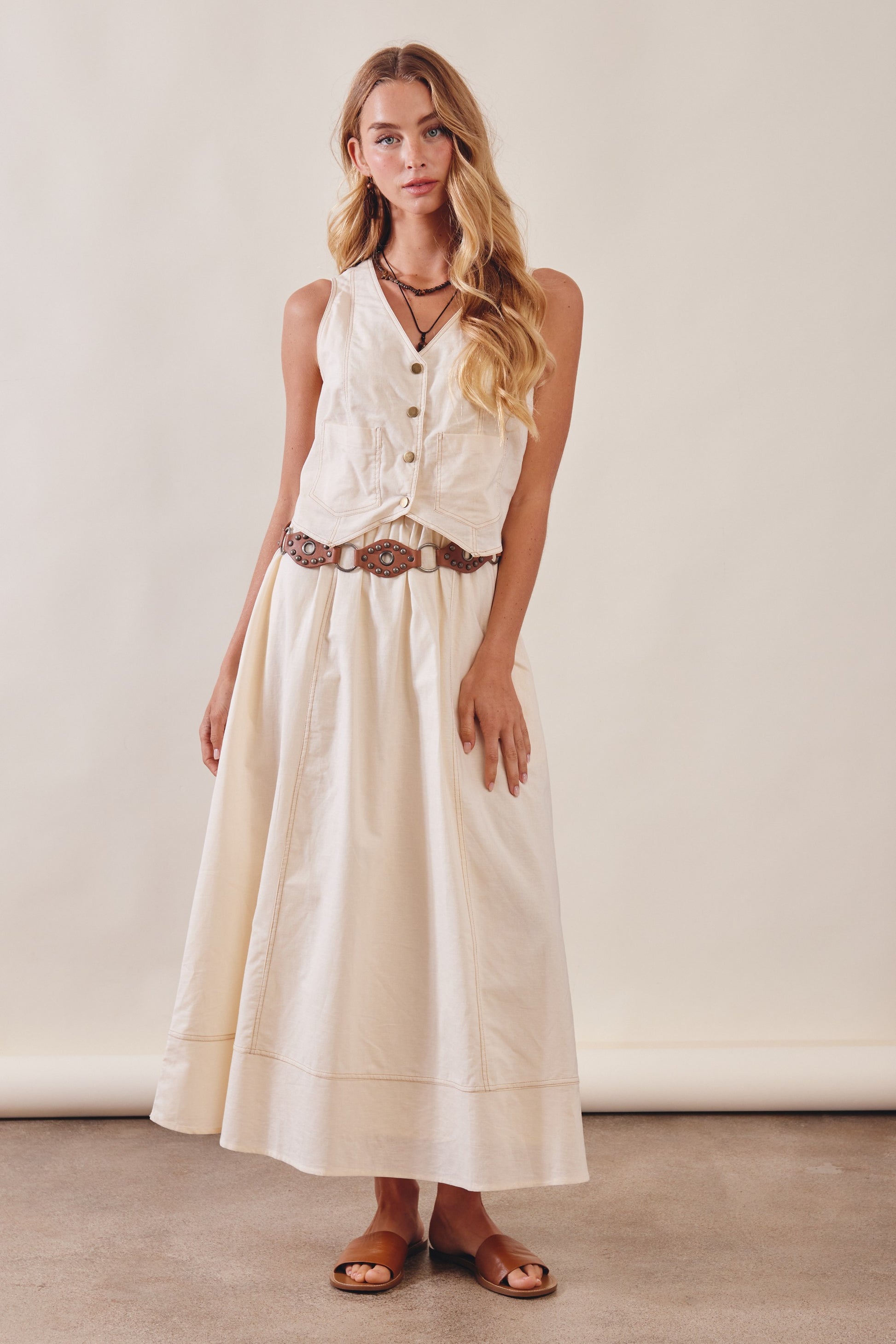Woman wearing a white sleeveless top and long beige skirt with a belt against a plain background