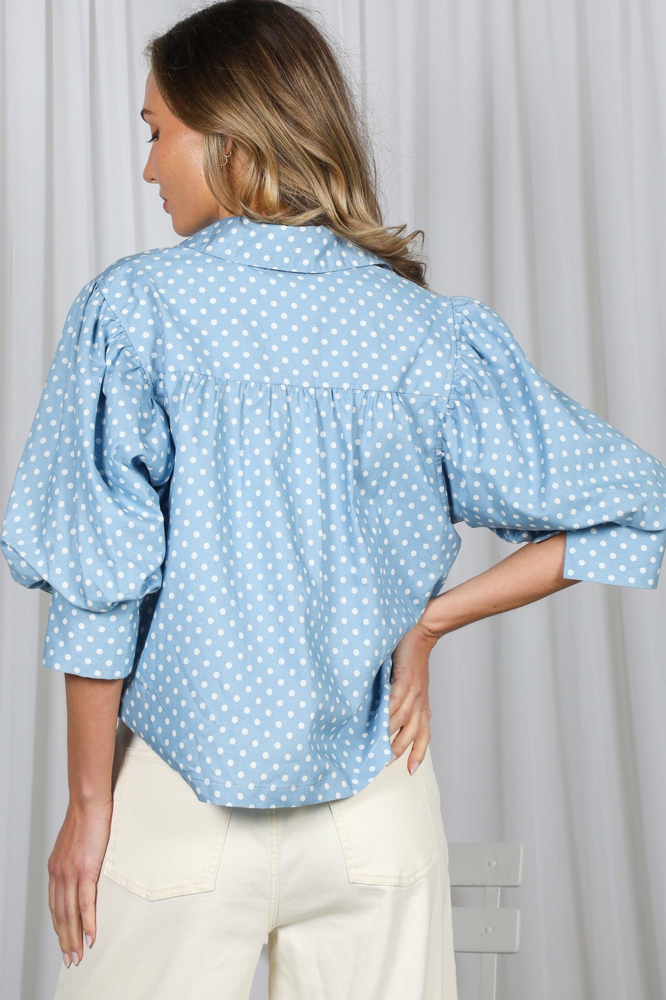 Woman wearing a light blue polka dot shirt with rolled-up sleeves against a white curtain background
