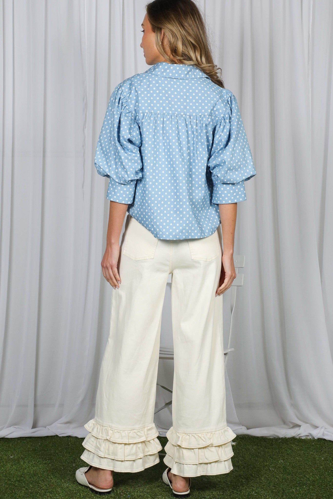 Woman wearing a blue polka dot blouse and white pants with ruffles in front of a white curtain.