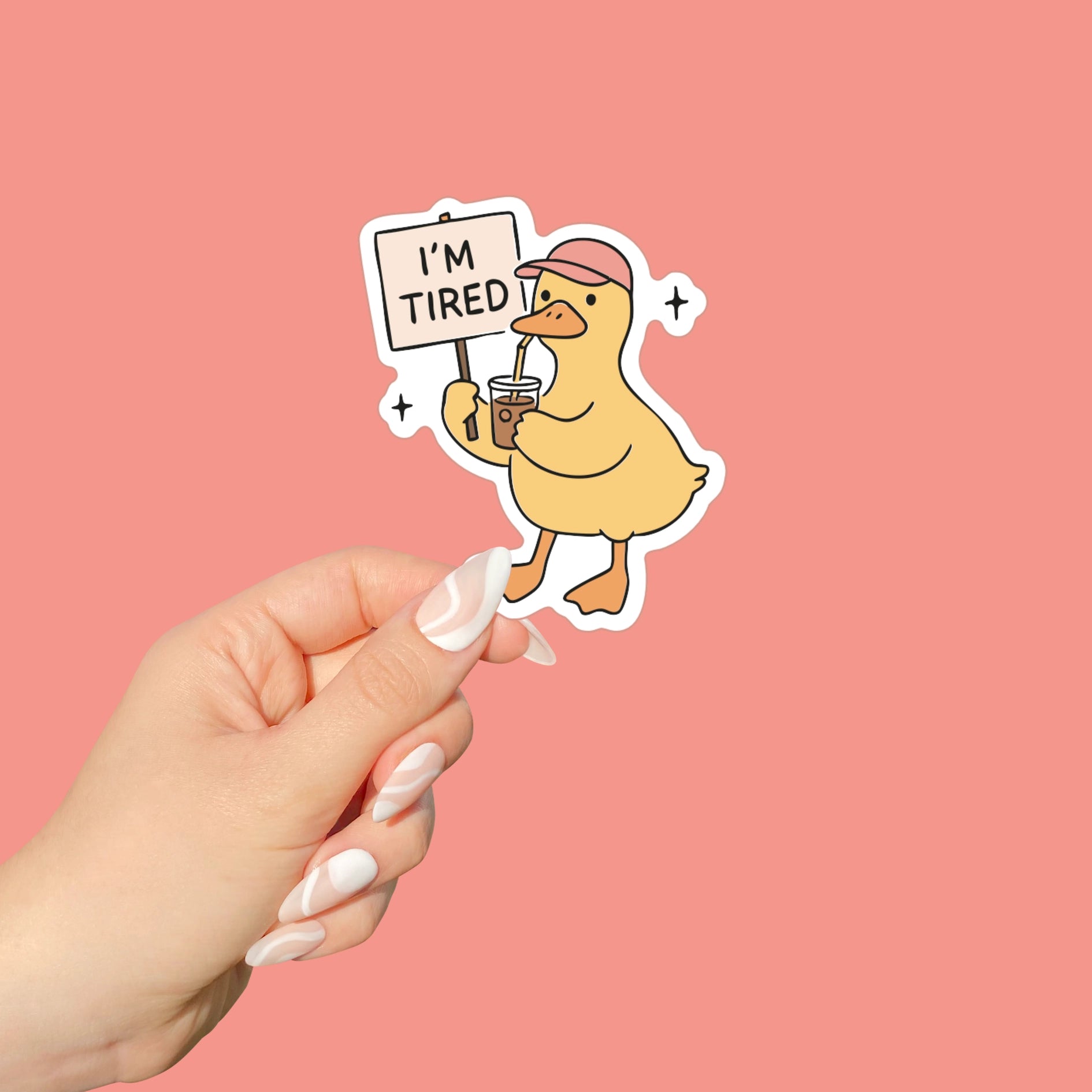 Hand holding a sticker of a duck holding a sign that says 'I'm Tired' on a pink background
