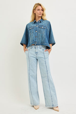 A blonde model wearing a cropped blue denim jacket with short sleeves and light-wash wide-leg denim jeans, standing against a plain off-white studio background.