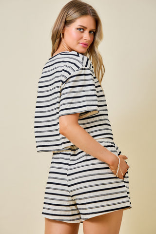 A woman modeling a matching short-sleeve top and shorts set with thin horizontal navy and off-white stripes, photographed against a plain beige studio background.