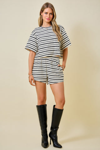 A woman modeling a matching two-piece set consisting of a short-sleeve top and shorts in a black and white horizontal stripe pattern, styled with black knee-high boots against a solid beige background.