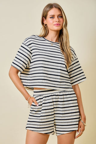 A woman modeling a matching two-piece loungewear set featuring a short-sleeve crop top and shorts, both with horizontal navy and cream stripes, set against a solid beige background.