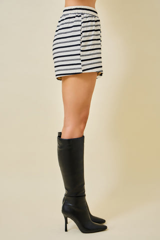 A side view of a model wearing a black and white horizontally striped mini skirt paired with black knee-high heeled boots, set against a solid beige studio background.