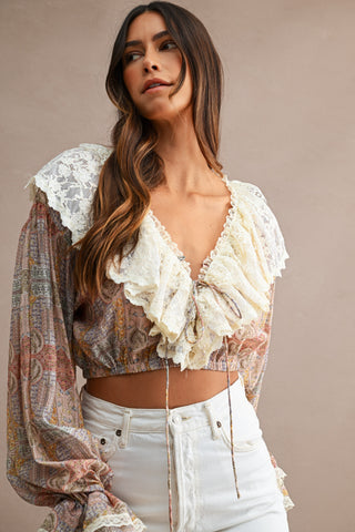 A woman wearing a bohemian-style long-sleeve crop top with a beige and brown paisley print, cream lace ruffle trim, and a tie-front neckline, paired with white jeans against a solid beige background.