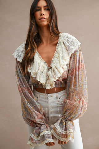 A woman wearing a bohemian-style sheer long-sleeve crop top with a cream lace ruffle collar and a multicolor paisley pattern, paired with white jeans, against a neutral beige background.