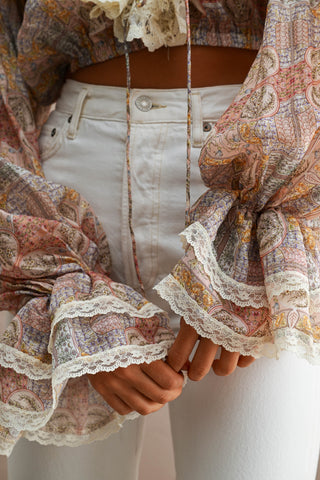 A close-up of a person wearing a bohemian paisley print top with dramatic ruffled sleeves and lace trim, paired with off-white jeans.
