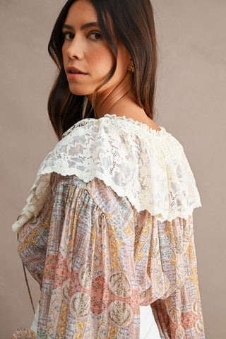 A woman wearing a bohemian-style long-sleeve blouse with a multicolor script print and a prominent white lace ruffle collar, photographed against a neutral beige background.