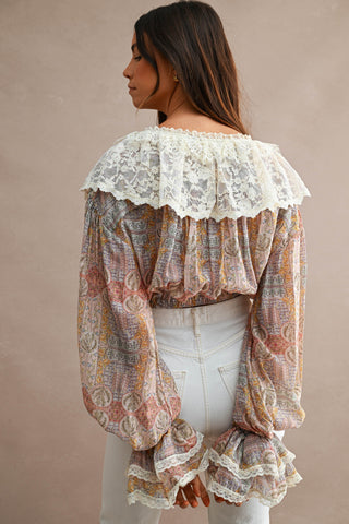 A woman shown from the back wearing a long-sleeve blouse with a cream lace ruffled collar and a multicolor paisley pattern, paired with white jeans against a plain beige studio background.