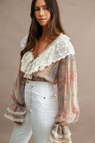 A woman wearing a bohemian long-sleeve blouse with a cream lace ruffle collar and cuffs, paired with white jeans, set against a neutral beige background.