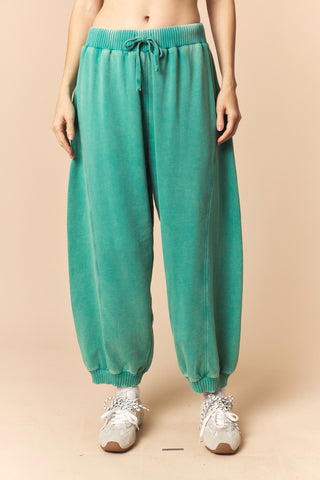 A person wearing loose teal-green drawstring lounge pants with elastic cuffs, paired with white sneakers, against a plain beige studio background.