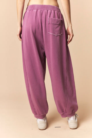 A person shown from the back wearing loose, wide-leg jogger-style pants in a dusty mauve-purple color, paired with white sneakers, against a solid beige studio background.