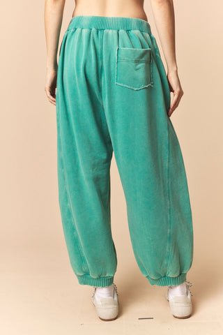 A person shown from the back wearing loose teal-green corduroy harem pants with an elastic waistband, back pocket, and ribbed ankle cuffs, paired with white sneakers against a plain beige studio background.