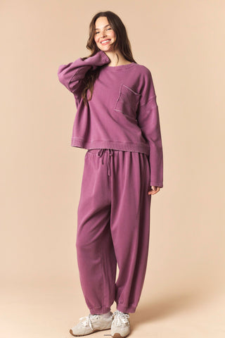 A woman modeling a matching two-piece lounge set in a dusty purple color, featuring a cropped long-sleeve knit top with a chest pocket and matching drawstring sweatpants, set against a solid beige background.