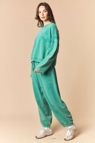 A woman modeling a matching two-piece lounge set in a vibrant teal green, featuring a cropped long-sleeve top with ribbed cuffs and matching loose jogger pants, paired with white sneakers against a solid beige background.