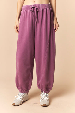 A person wearing loose, wide-leg jogger sweatpants in a dusty mauve-purple color, paired with white sneakers, against a solid beige studio background.