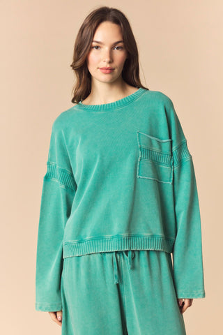 A woman modeling a matching two-piece loungewear set in a washed teal-green color, featuring a cropped long-sleeve top with a chest pocket and matching drawstring pants, set against a solid beige background.