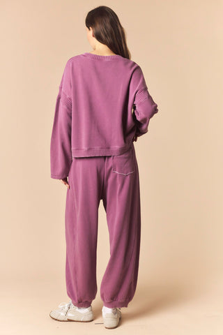 A woman shown from the back wearing a matching mauve-purple cropped sweatshirt and wide-leg sweatpants set, paired with white sneakers, against a solid beige studio background.