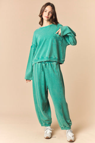 A woman modeling a matching two-piece lounge set in a washed teal-green color, featuring a cropped long-sleeve sweatshirt with a chest pocket and matching jogger pants, paired with white sneakers against a solid beige background.