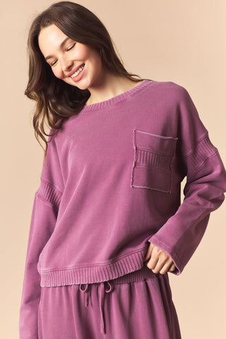 A woman wearing a dusty purple knit sweater with a chest pocket and matching drawstring pants, posed against a solid beige background.