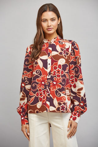 A woman modeling a long-sleeve button-up blouse with a bold retro floral and swirl pattern in red, orange, brown, and cream, paired with cream-colored pants against a neutral gray background.