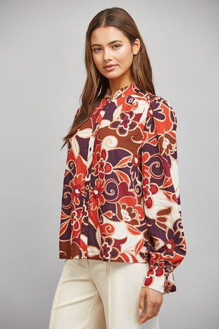 A woman modeling a long-sleeve blouse with a bold retro floral and swirl pattern in rust red, orange, brown, and cream, paired with cream pants against a solid gray studio background.