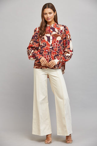 A woman modeling a long-sleeve blouse with a red, orange, and brown floral and paisley pattern, paired with wide-leg cream trousers, against a solid gray studio background.