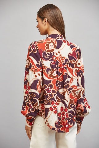 A woman shown from the back wearing a long-sleeve blouse with a bold floral and paisley-style print in rust red, cream, and dark purple, paired with cream pants, against a neutral gray studio background.
