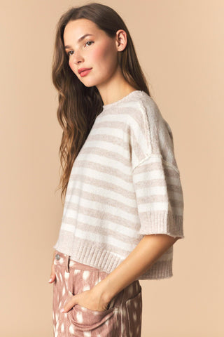 A woman wearing a beige and white horizontally striped knit sweater, paired with pink patterned pants, set against a solid beige background.