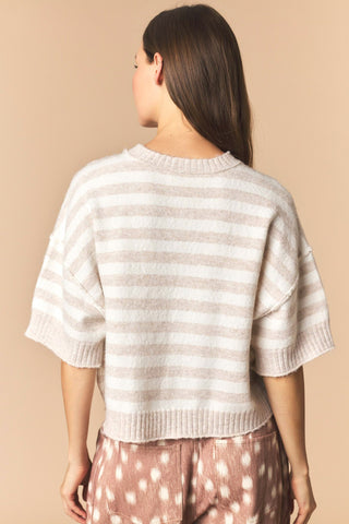 A woman shown from the back wearing a short-sleeve knit sweater with horizontal beige and off-white stripes, paired with pink patterned pants, against a solid beige studio background.